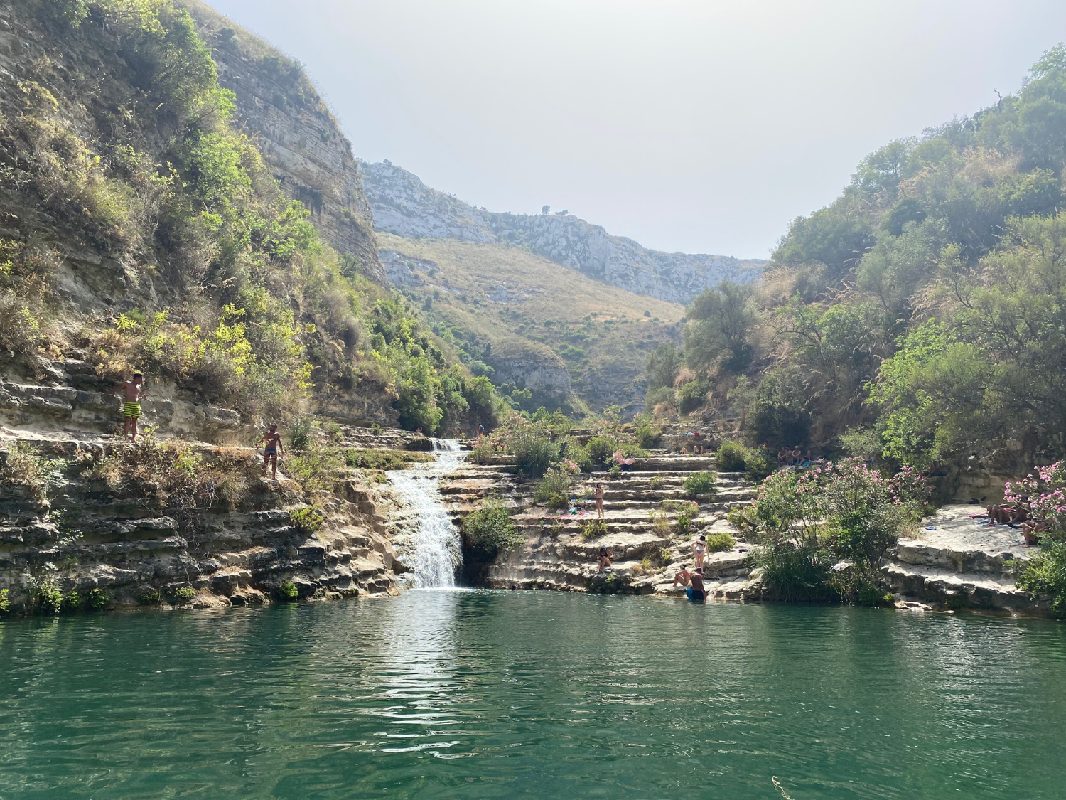 Scenic view of the natural reserve Cavagrande del Cassibile in Sicily, featuring a clear, emerald green pool with cascading waterfalls surrounded by lush vegetation and rocky cliffs