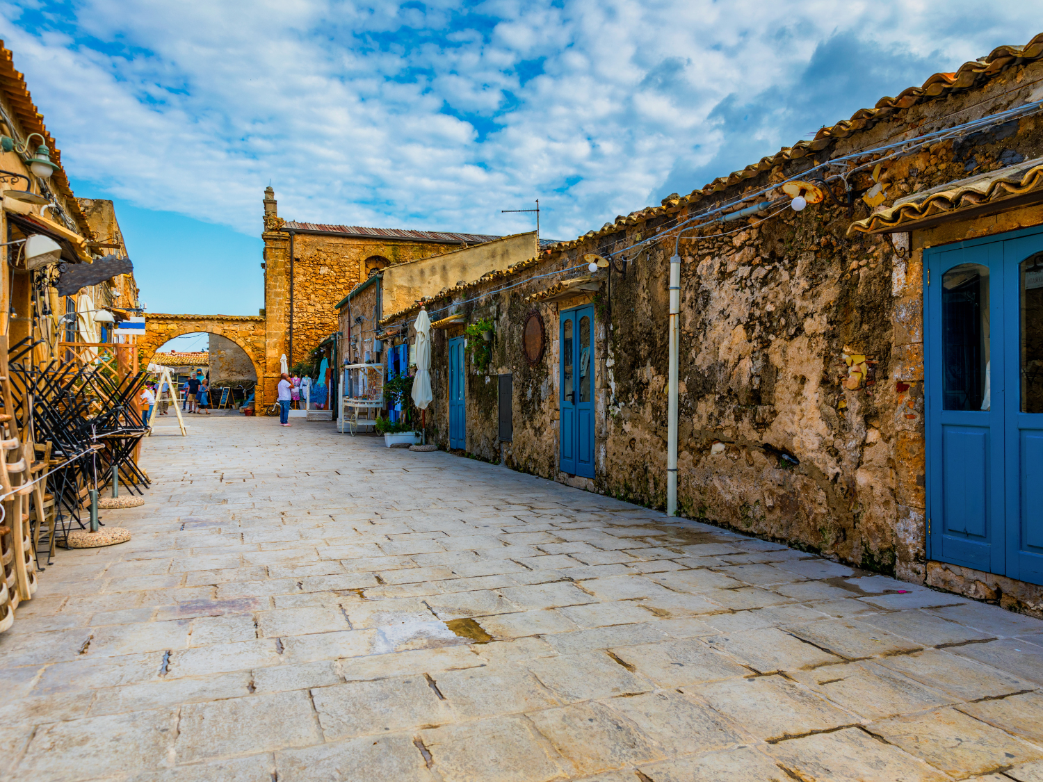 A charming stone-paved street in Marzamemi, Sicily, with rustic buildings, blue doors, and a scenic archway under a bright blue sky.